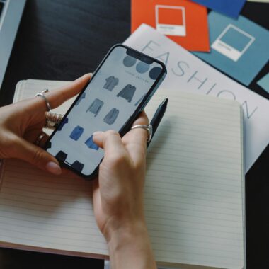 A woman browsing fashion items on a smartphone while sitting at a desk indoors.